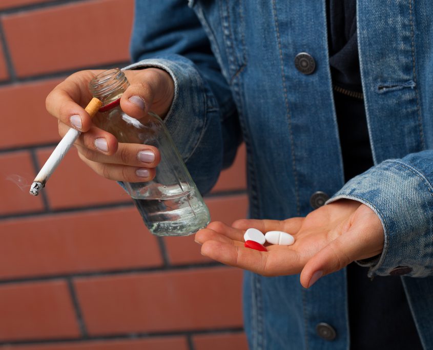 A closeup of hand holding a cigarette, vodka and pills